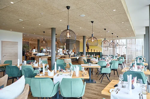 A modern, bright dining area at a vineyard, featuring set wooden tables and mint-green upholstered chairs, wire pendant lights, a wine barrel bar, a large glass front on the right and a wooden door on the left.