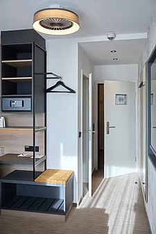 Hotel-Eingangsbereich: geöffnete weiße Zimmertür mit Griff, schmaler Gang; links offene Garderoben-/Regallösung in Holz und schwarzem Metall mit Safe, Haken, Sitzbank, Deckenleuchte und Wandspiegel.