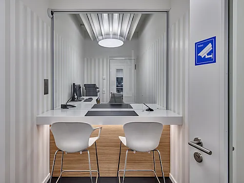 Raum mit Servicetresen hinter einer gläsernen Schutzscheibe: weiße Theke mit zwei hohen weißen Stühlen, dahinter Computerarbeitsplatz, gestreifte Wände, runde Deckenleuchte, Videoüberwachungssymbol am Türrahmen.