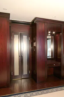Porte intérieure à deux battants en bois foncé, avec vitrage satiné et poignées rondes en métal, encastrée dans un lambris mural couleur acajou ; à droite, une niche avec miroir, tiroir et applique murale ; parquet foncé bordé d'une moquette.