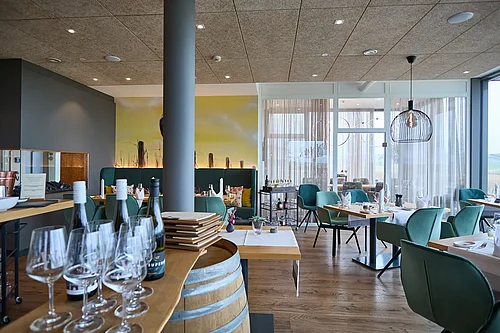 A bright dining room at a vineyard with wooden flooring: tables laid with tablecloths, green upholstered chairs, bottles of wine and wine glasses on a wooden barrel in the foreground; the room is divided into sections by a transparent sliding glass door/glass partition with curtains; ceiling lights and a column are visible.