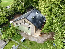 Ripresa da drone di una piccola casa con tetto a falde scure e due lucernari, facciata chiara con rivestimenti in legno verde, finestre strette, passerella in legno laterale come ingresso, circondata da alberi