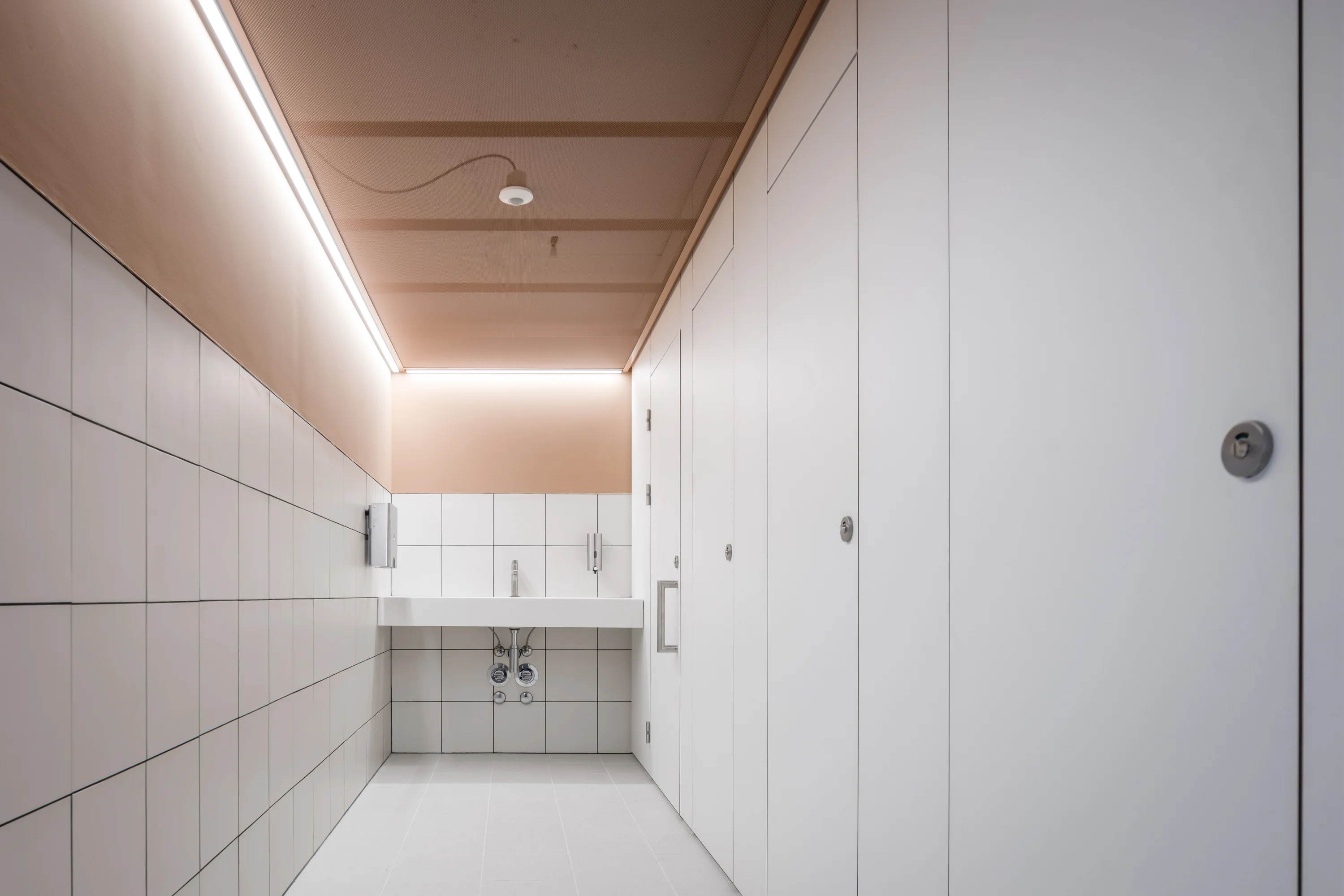 Heller, minimalistisch gestalteter Sanitärraum: langes weißes Wandwaschbecken mit zwei Armaturen und freigelegten Leitungen, links geflieste Wand, rechts mehrere geschlossene weiße Kabinentüren mit runden Schlössern; helle Holzdecke mit durchgehendem LED-Band.