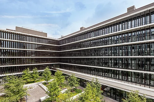 Vue extérieure du bâtiment de laboratoires en forme de U de l'université de Zurich (Irchel), doté d'une façade vitrée à plusieurs étages et de lamelles verticales, de niveaux/balcons sur tout le pourtour, ainsi que d'une cour intérieure végétalisée agrémentée d'arbres et d'espaces de détente.