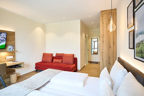 A bright guest room with a double bed in the foreground, white cushions and a white duvet, a red sofa bed against the left-hand wall, light-coloured wooden panelling with a small desk and TV, a hanging bulb pendant light on the right, and an open interior door leading to a short corridor with a mirror.