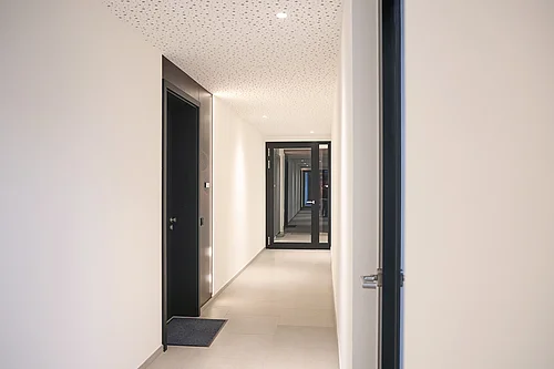 Luminoso corridoio interno con pareti bianche e piastrelle al pavimento; a destra e a sinistra, strette porte interne scure con maniglie metalliche e telai. In fondo, un’ampia porta a vetri con cornice nera che prolunga il lungo corridoio illuminato. Davanti a una porta c’è uno zerbino scuro.