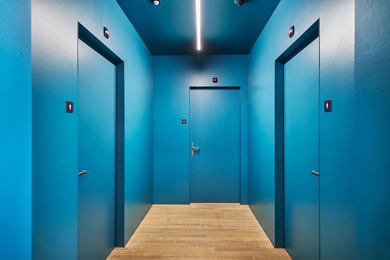 Ripresa interna di uno stretto corridoio con pareti blu brillante e tre porte uniformemente blu. Porta centrale con maniglia, porta destra e sinistra con pittogrammi di WC, pavimento in legno chiaro, plafoniera lineare.