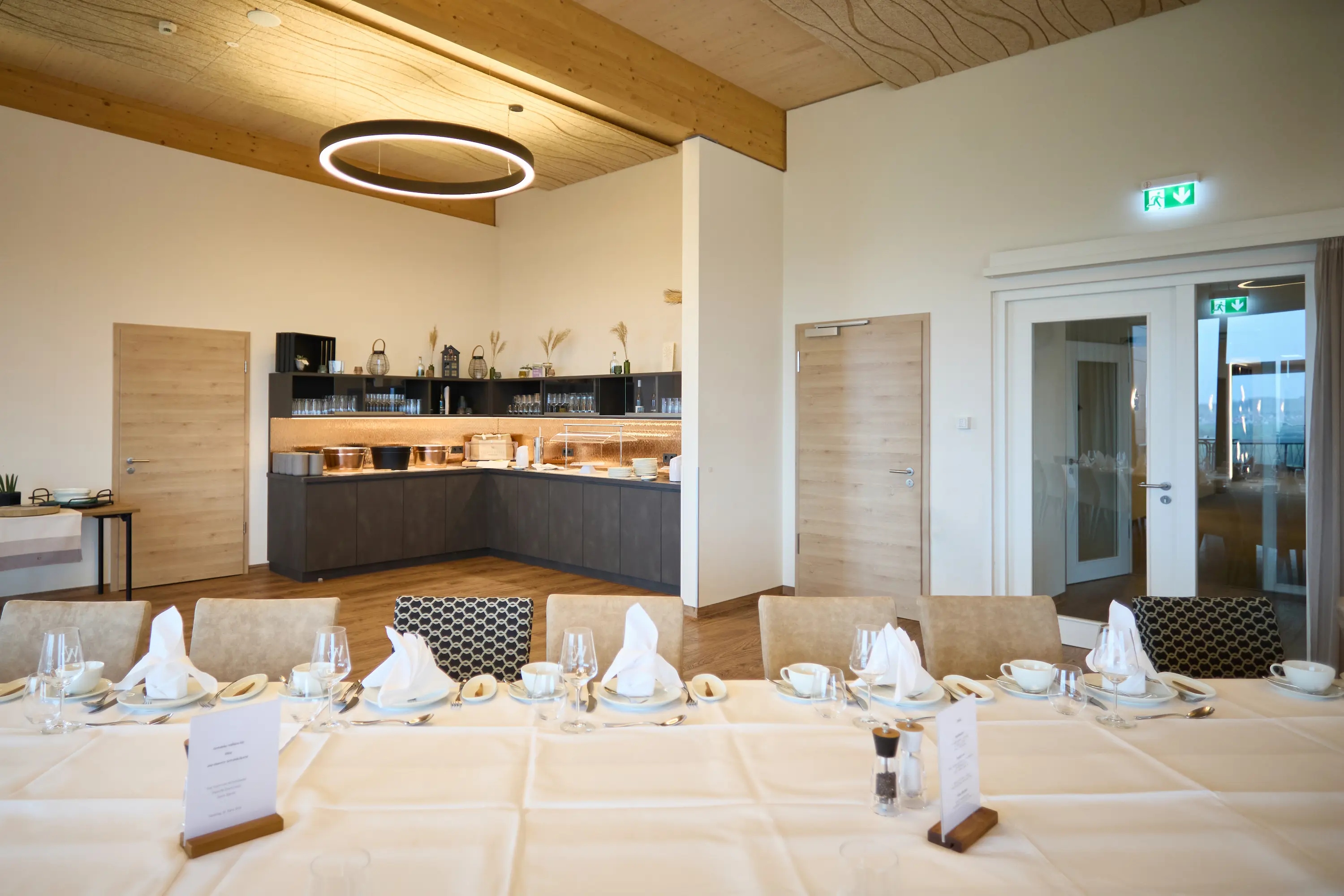 A wide-angle shot of a laid banquet table in a brightly lit dining room: a long white tablecloth with plates, glasses, cutlery and folded napkins. In the background, a dark buffet counter, two light-coloured wooden interior doors and, on the right, a pair of white glass double doors with an illuminated emergency exit. The room features wooden parquet flooring, an exposed wooden ceiling and a round pendant light, creating a warm, modern atmosphere.