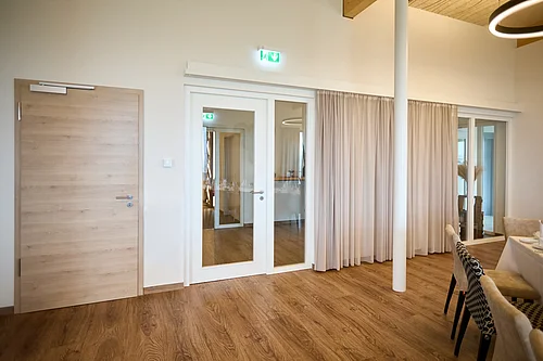 A bright room with wooden flooring: on the left, a solid wooden door with a door closer; in the centre, a white double-leaf glass door with an emergency exit sign; on the right, a curtain in front of a sliding glass door; a pillar and a laid table are visible.