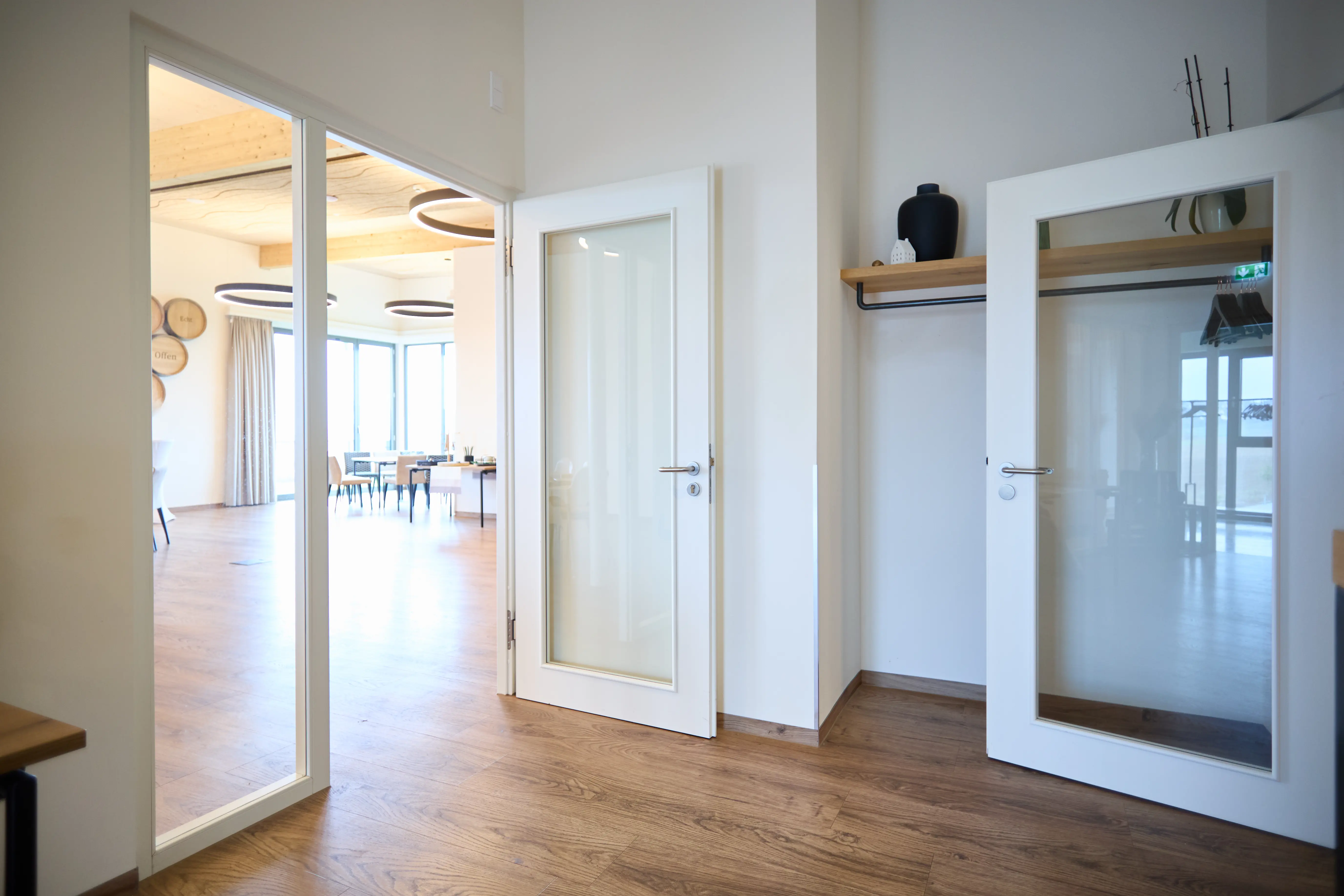 A bright entrance hall with wooden flooring: two white interior doors with large glass panels; on the left, an open door with a glass side panel leading to the bright lounge/dining room with large windows and round ceiling lights; on the right, a recess with a wooden shelf and a vase.