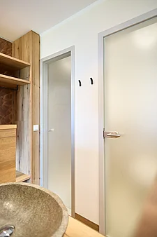 Hallway area with two frosted glass doors in light grey frames (door on the right with a metal handle, narrow passageway on the left); wooden shelf on the left, stone washbasin at the front and two black wall hooks