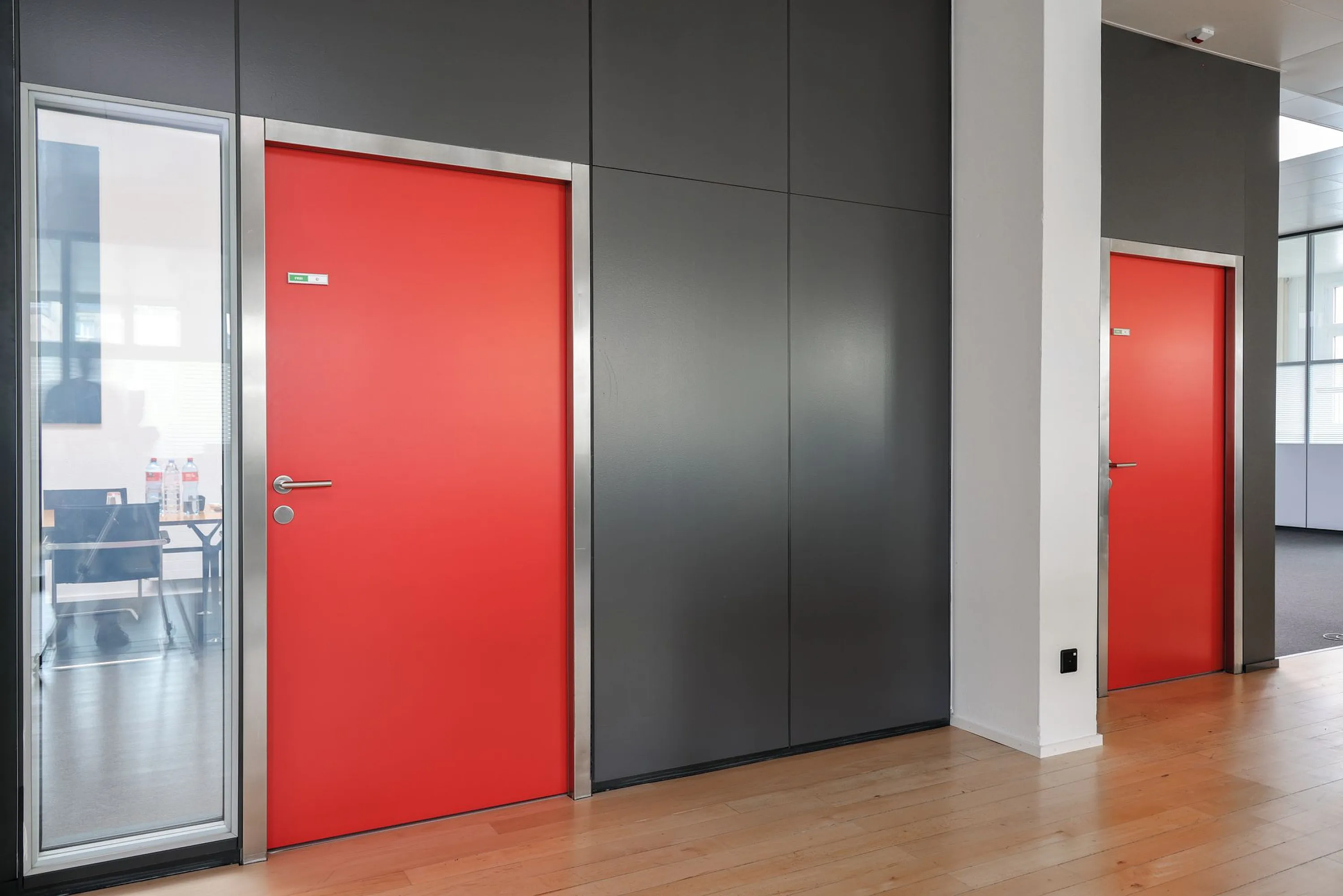 Blick auf zwei mattrote Innentüren mit silberner Metallzarge und Hebelgriffen in einer dunklen Paneelwand; links schmale Glas-Trennwand zum Meetingraum, heller Holzfußboden.