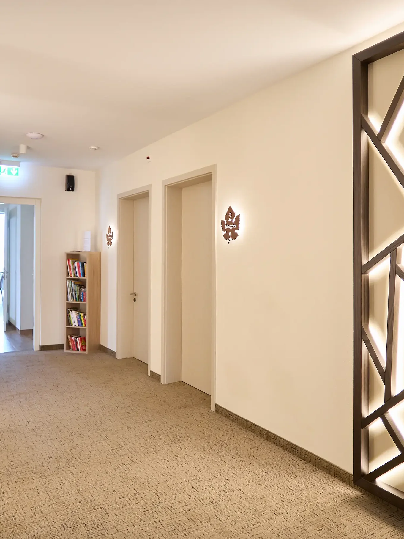 Bright corridor in the guest house/winery: beige carpet, two solid interior doors on the right, a double-leaf glass door/sliding glass door at the end, a large backlit geometric wooden panel on the right, a small bookcase and an emergency exit sign.