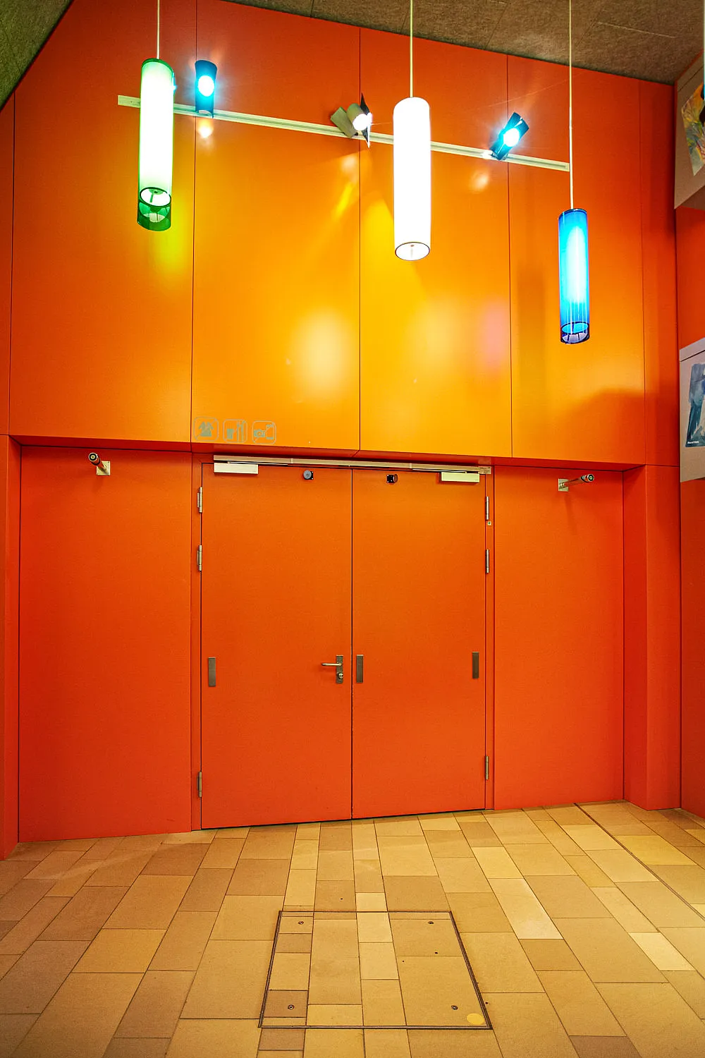 Porta a due battenti di colore arancione in un rivestimento continuo di colore arancione; tre luci cilindriche a sospensione di colore verde, bianco e blu al di sopra; pavimento in piastrelle di colore chiaro; maniglia, chiudiporta e due piccoli pensili sopra la porta.