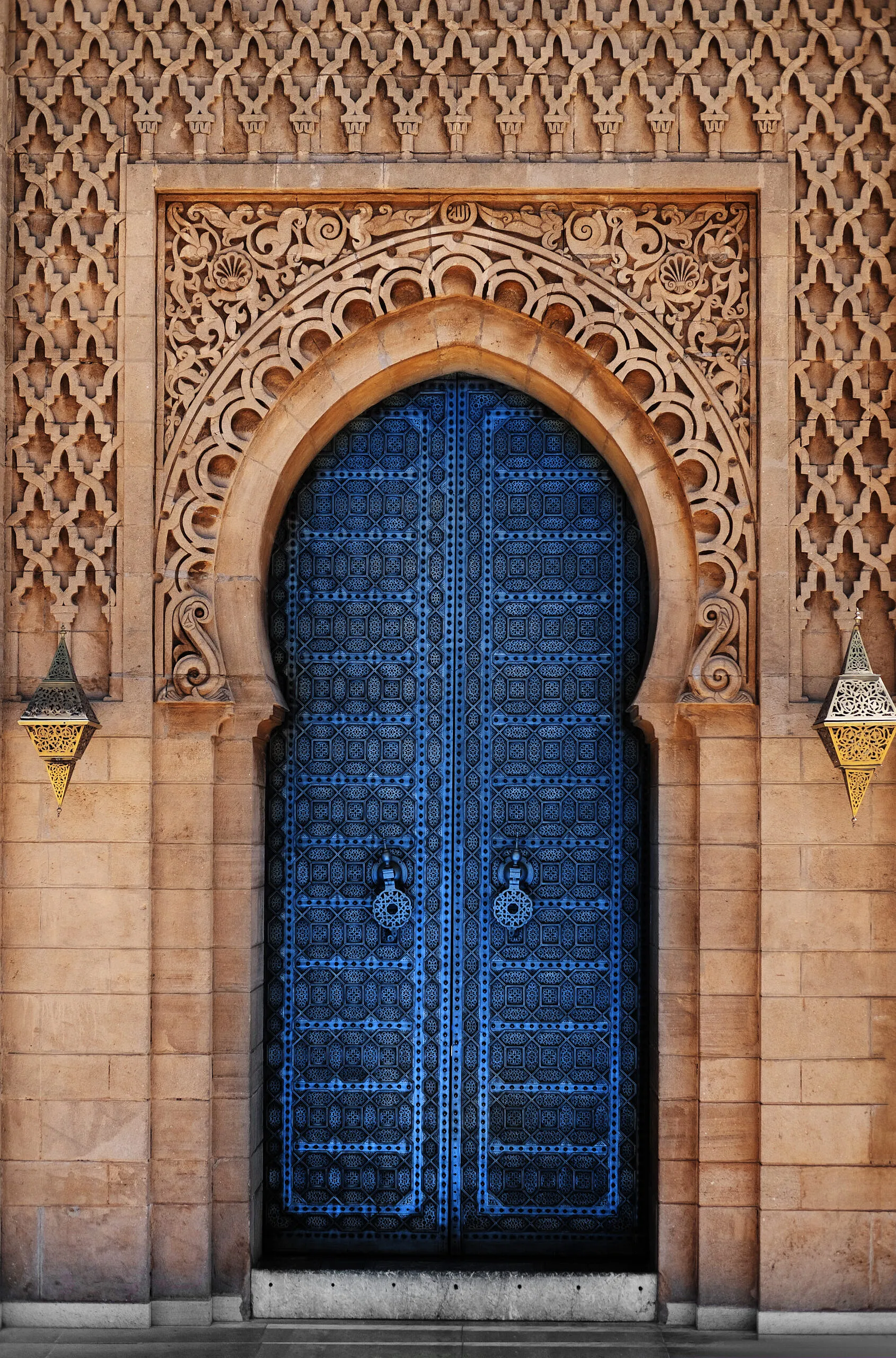 Haute porte à double battant d'un bleu profond, ornée de motifs géométriques fins et de heurtoirs ronds, encadrée d'un arc en pierre sculpté avec des ornements arabesques et deux lanternes