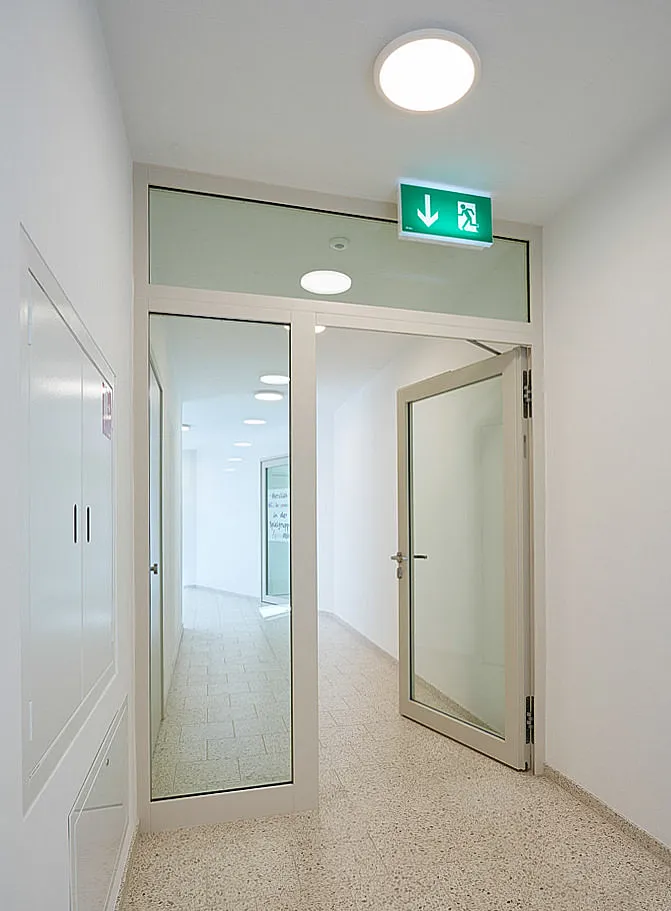 Innenflur mit geöffneter einflügeliger Glastür in hellem Metallrahmen und festem Seitenteil, Oberlicht, grünes Notausgangsschild mit Laufmann und Pfeil nach unten, runde Deckenleuchten und heller Terrazzo-Boden.