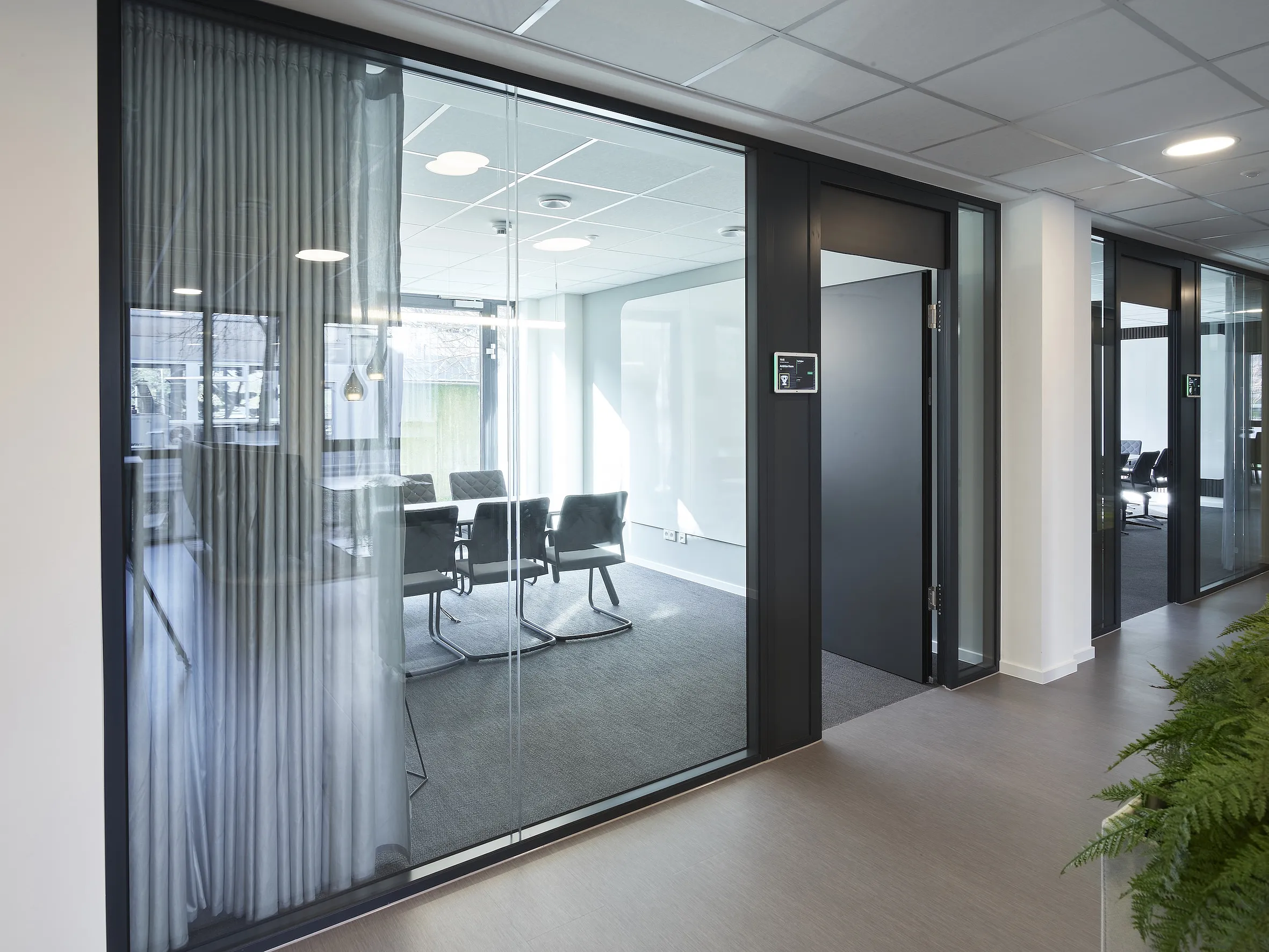 Glasraumtrenner mit schwarzem Metallrahmen und halbtransparentem Vorhang; dahinter Besprechungsraum mit Tisch und mehreren Stühlen, Teppichboden und Rasterdecke mit Einbauleuchten; geöffnete dunkle Tür mit elektronischem Türschild; Flur mit Pflanze