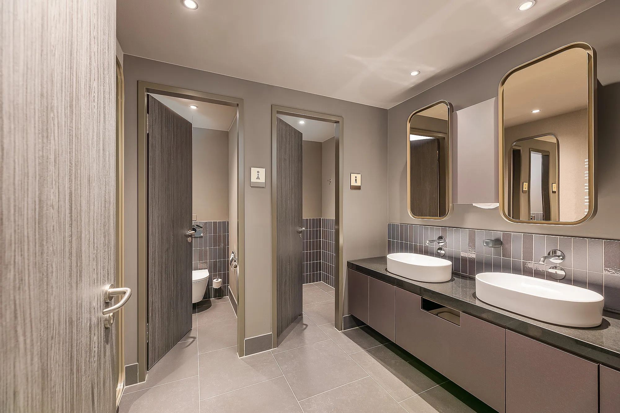 A modern, multi-user toilet block featuring two oval countertop washbasins set on a dark worktop, two rounded wall-mounted mirrors, grey floor and wall tiles, and three open-plan toilet cubicles with wood-effect doors.