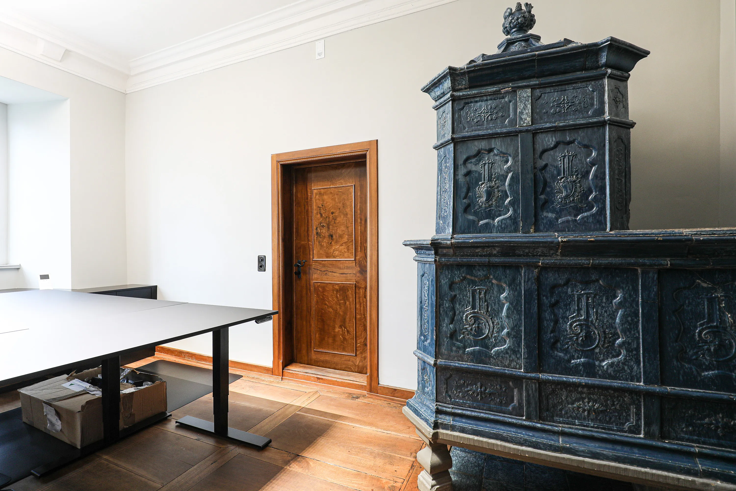 Pièce dans château : porte en bois brun avec huisserie et poignée noire, à droite un grand poêle en faïence sombre et orné, à gauche une fenêtre et une table de travail moderne avec des cartons en dessous.