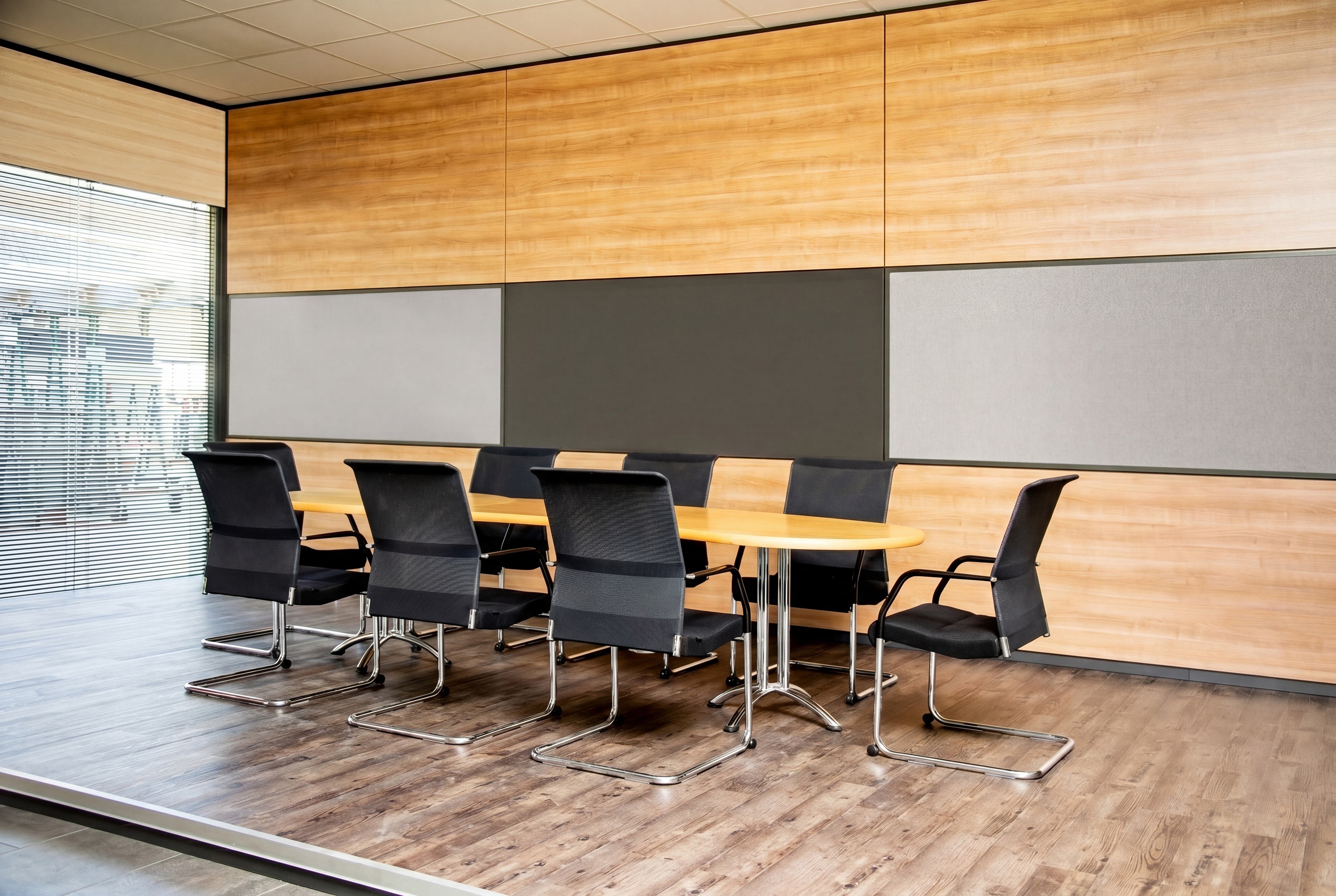 Sala riunioni con tavolo ovale in legno e diverse sedie nere con pattini in metallo; parete rivestita con pannelli in legno chiaro e pannelli acustici o divisori grigi; grandi finestre con tapparelle orizzontali sulla sinistra; pavimento in laminato, soffitto con pannelli a griglia.