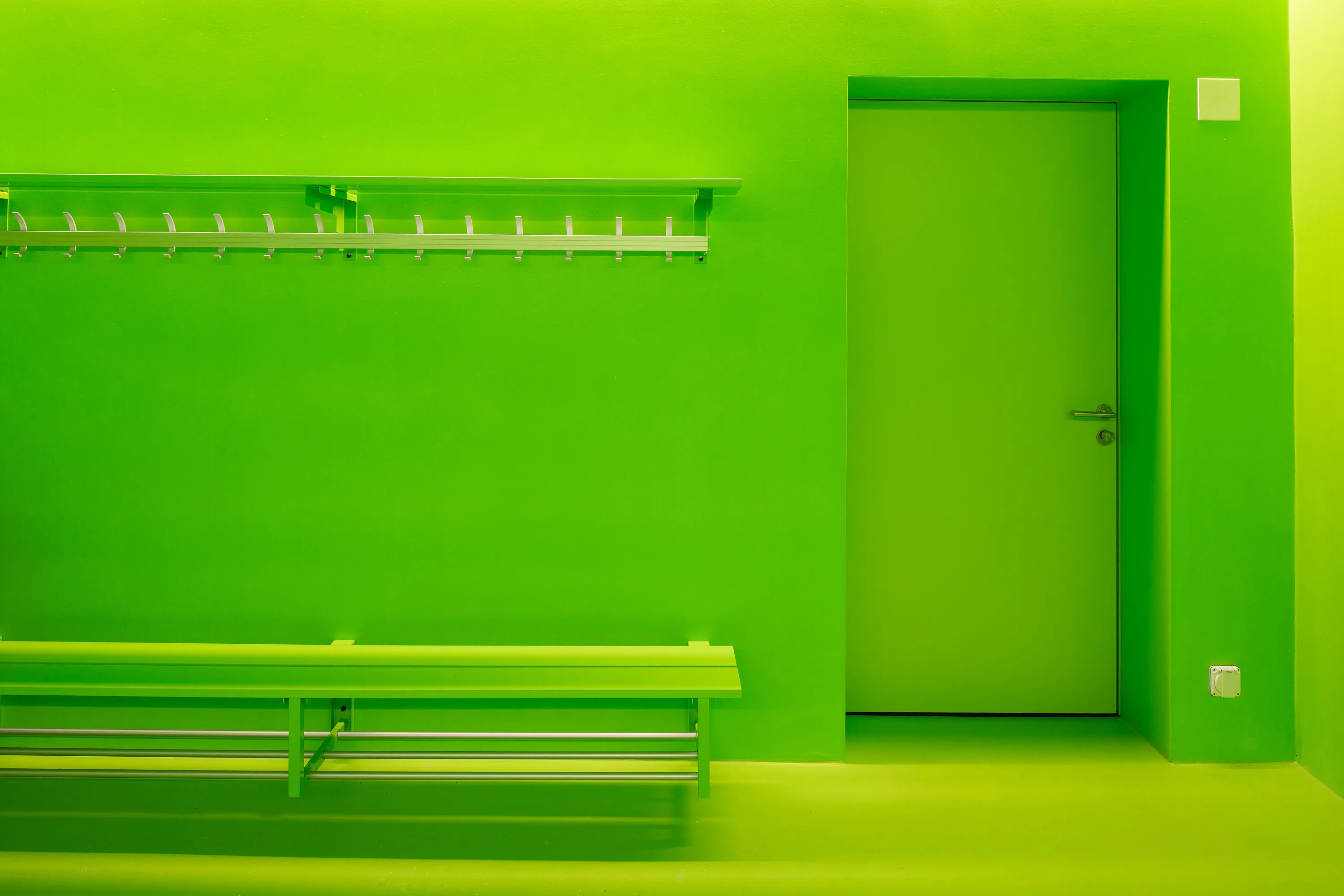 Vestiaire vert vif : sur le mur de droite, une porte simple et fermée avec poignée ; devant, un long banc et au-dessus, un vestiaire métallique avec plusieurs crochets. Une conception claire et réduite.