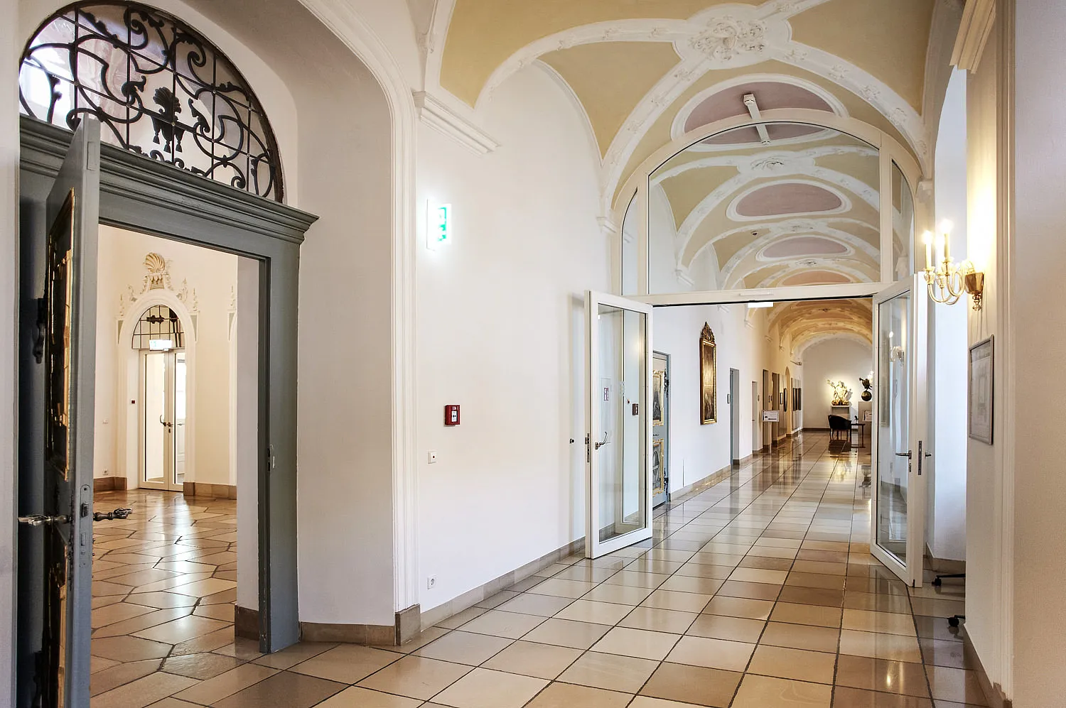 Lungo e luminoso corridoio interno con soffitto a volta in stucco dai colori pastello, pavimento in piastrelle quadrate lucide e diverse porte a vetri aperte; a sinistra un alto architrave con ferro battuto decorativo, a destra applique e quadri incorniciati, mobili a vista in fondo al corridoio.