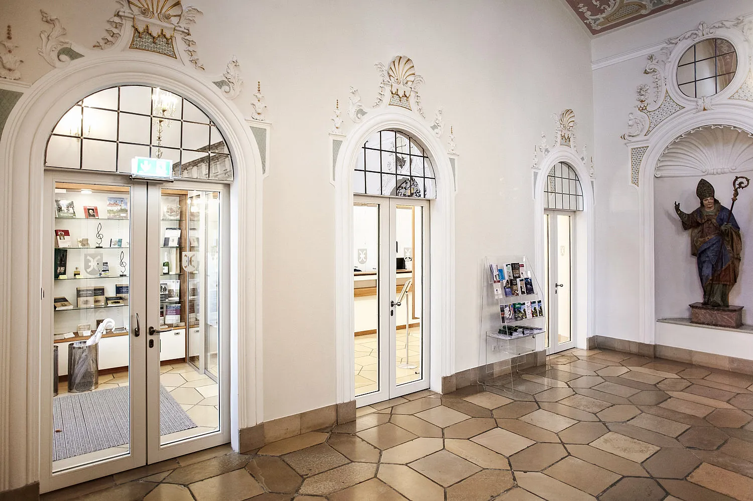 Interno con tre porte a vetri bianchi in cornici ad arco in stucco e sopraluce; cartello di uscita di emergenza e vetrine a sinistra, statua vescovile in nicchia a destra, portadepliant, pavimento in pietra naturale.