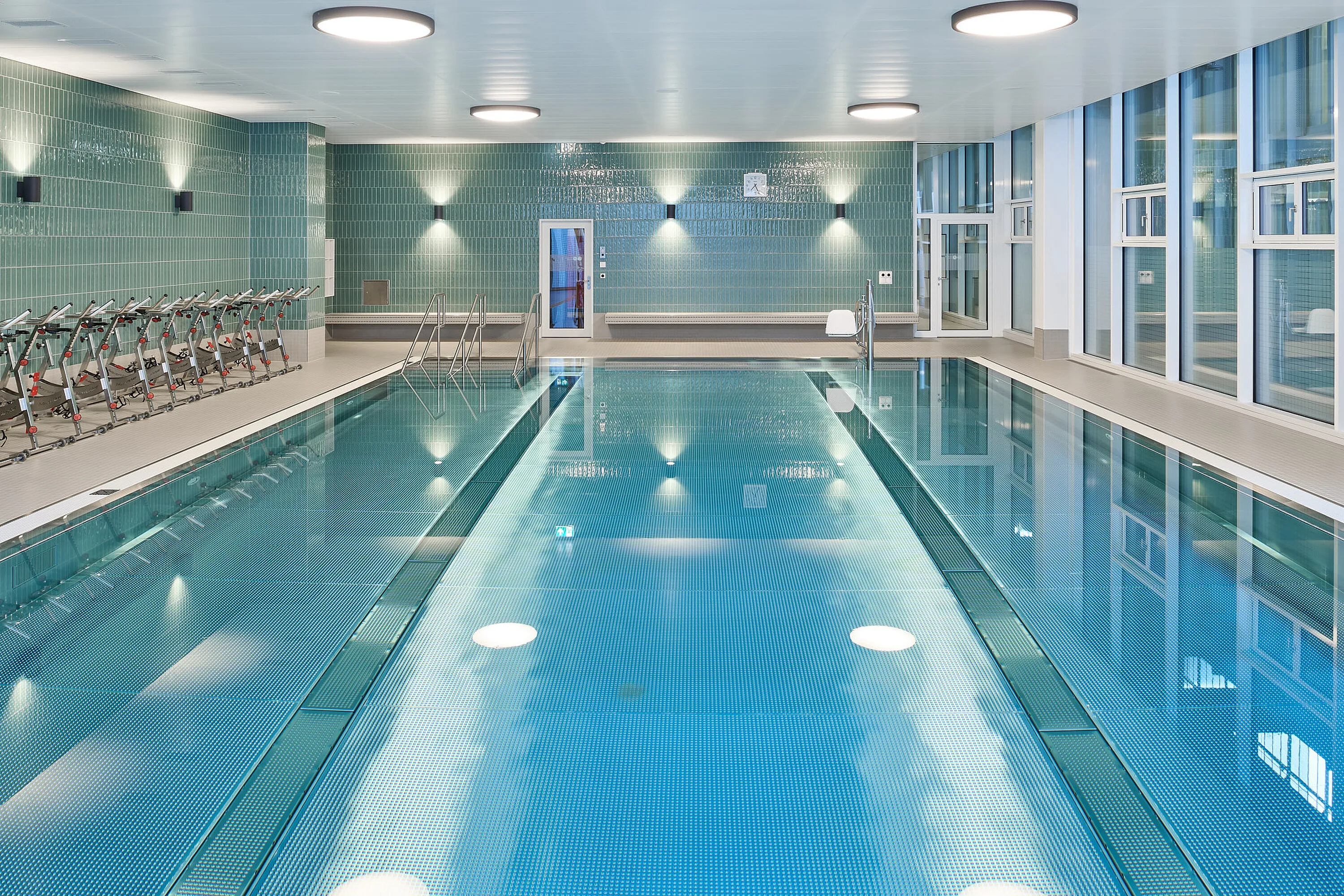 Piscina terapeutica coperta: vasca terapeutica rettangolare con gradini e corrimano, ascensore per piscina, fila di biciclette terapeutiche subacquee sul lato, piastrelle verdi a parete e grande finestra frontale.