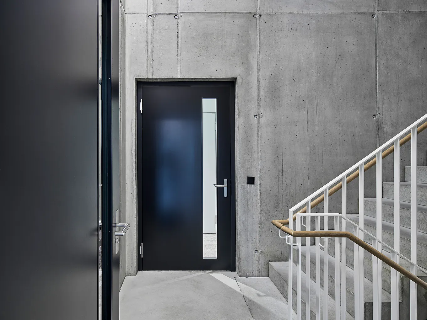 Vue intérieure de la cage d'escalier : porte gris foncé avec un étroit insert vertical en verre à droite et poignée en acier inoxydable, entourée de murs en béton apparent ; à droite, escalier avec rampe blanche et main courante en bois.