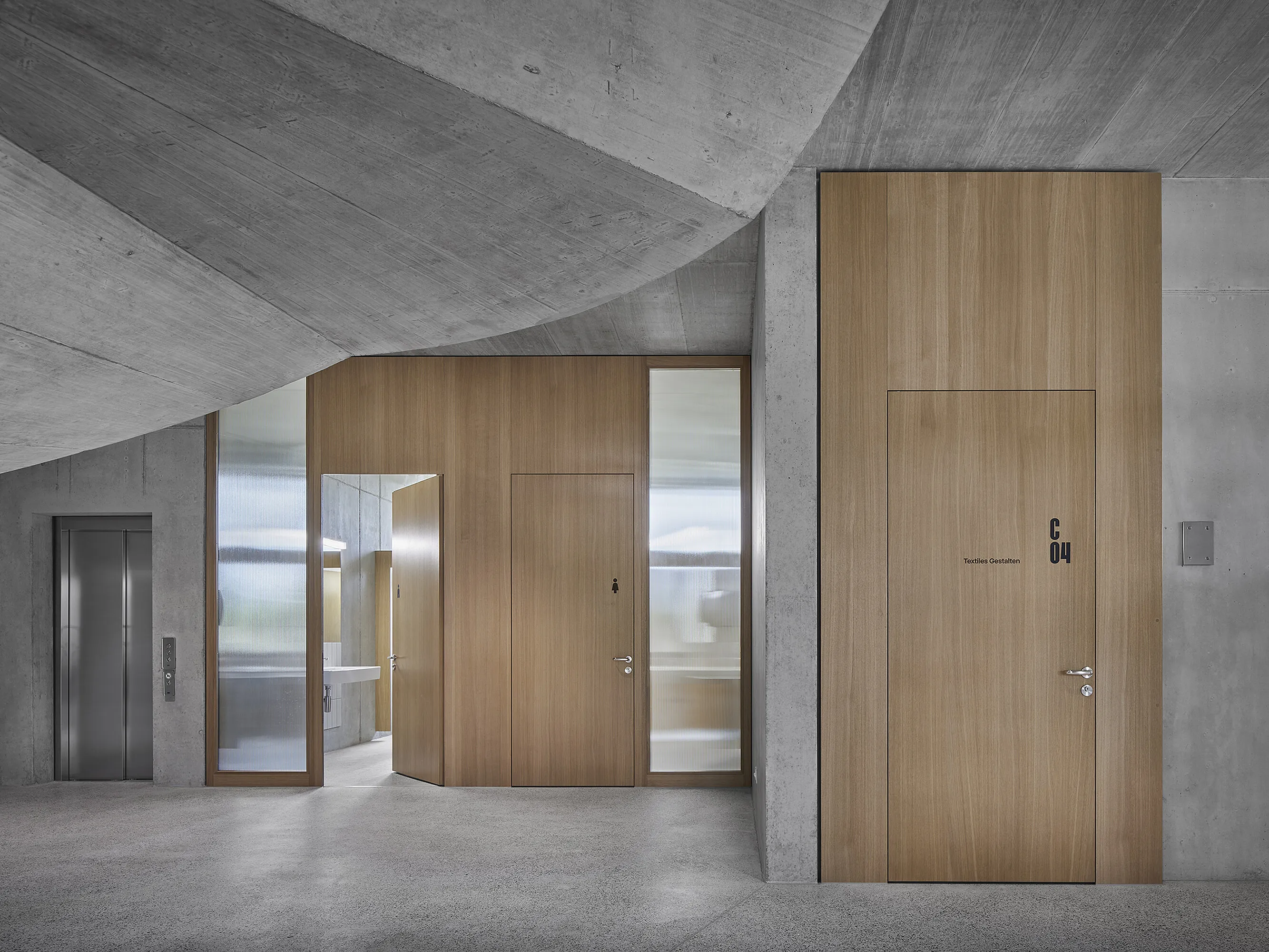 School corridor with exposed concrete ceiling: three wooden-clad interior doors in a wooden panel wall, a large door labelled 'C 04', an open passageway with frosted glass on the side and a lift on the left.