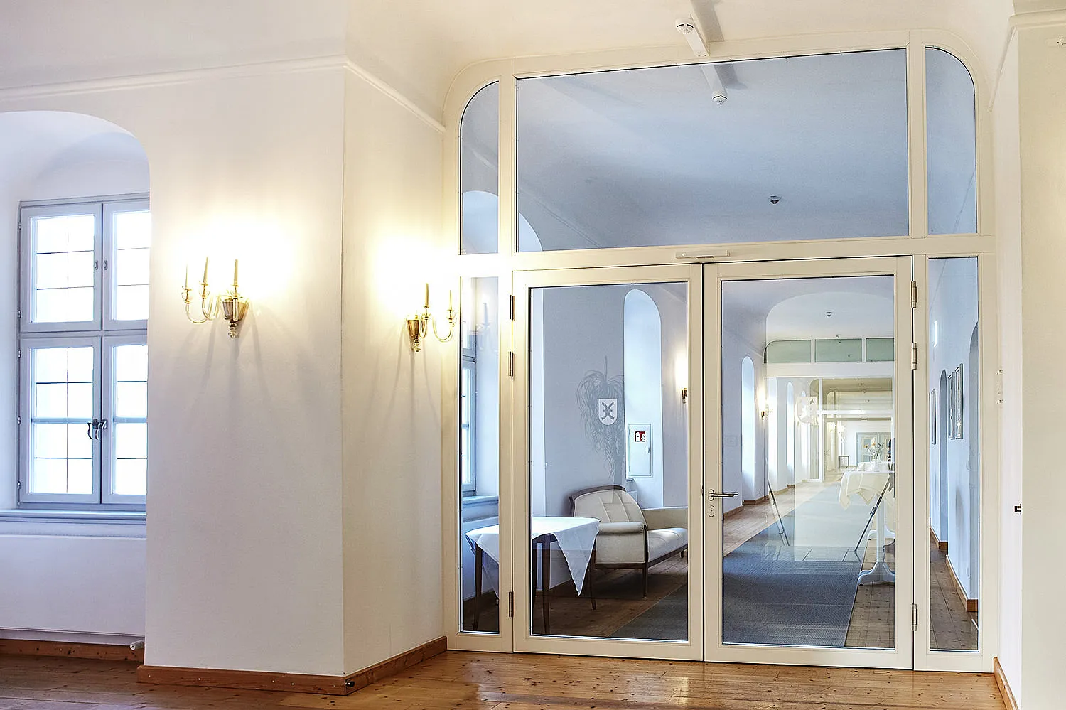 Porta in vetro con cornice bianca e sopraluce in corridoio chiaro; zona soggiorno con divano, tavolo, pavimento in legno e finestra a traforo visibile attraverso la porta