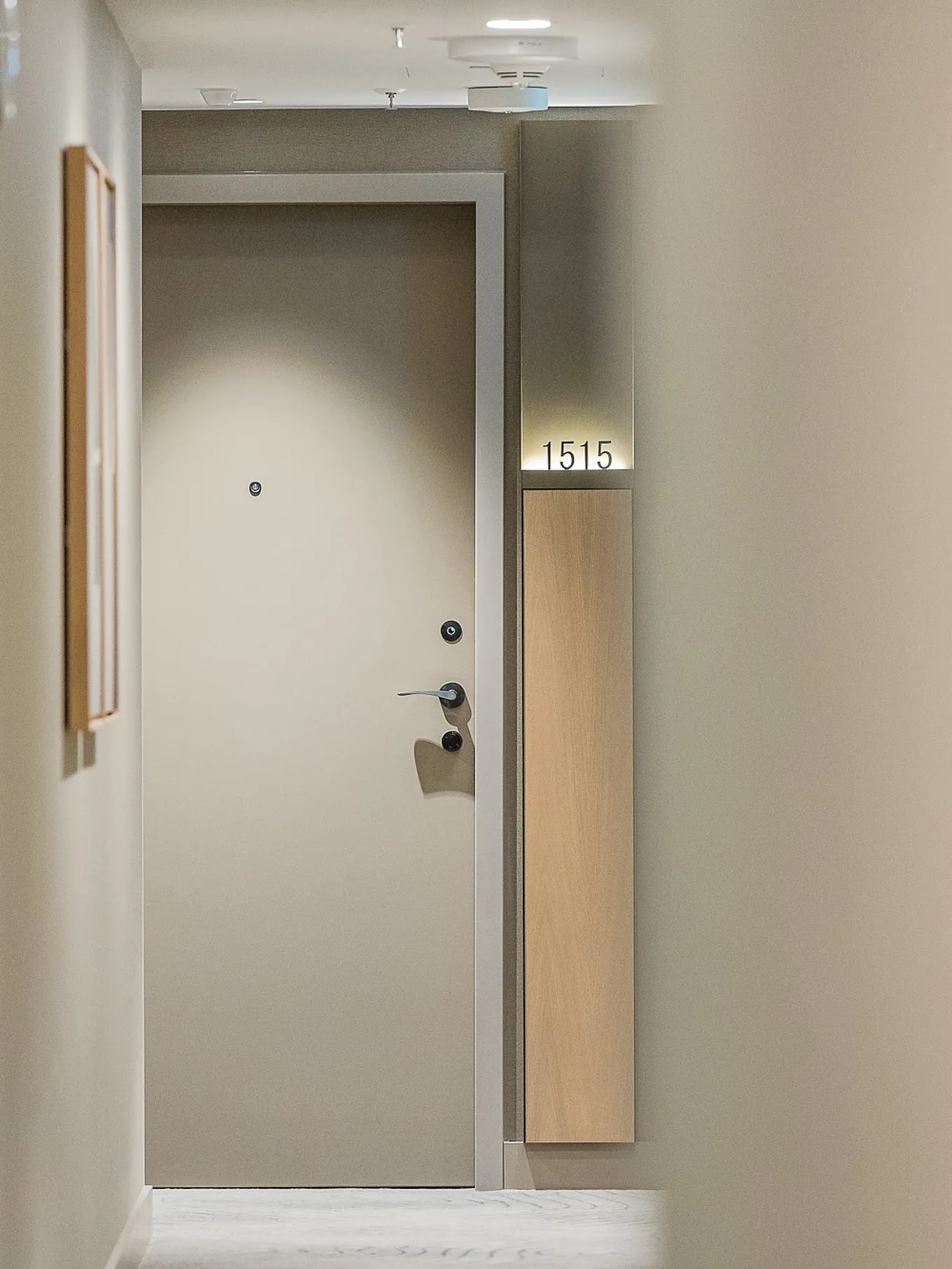 A narrow, brightly lit corridor; at the end, a solid, plain-coloured door with a spy and a handle; on the right, an illuminated door panel bearing the number 1515; on the left, blurred framed images on the wall.