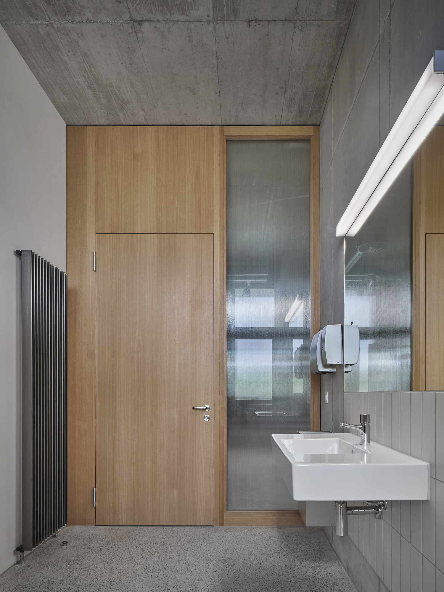 Vista interna di un bagno scolastico: porta interna in legno incassata a filo con telaio in legno a sinistra di un alto pannello laterale in vetro smerigliato. A destra, lavabo bianco sospeso con rubinetto, grande specchio e distributore di sapone in acciaio inox; a sinistra, radiatore nero sottile. Soffitto in cemento a vista, pavimento grigio e materiali sobri e chiari.