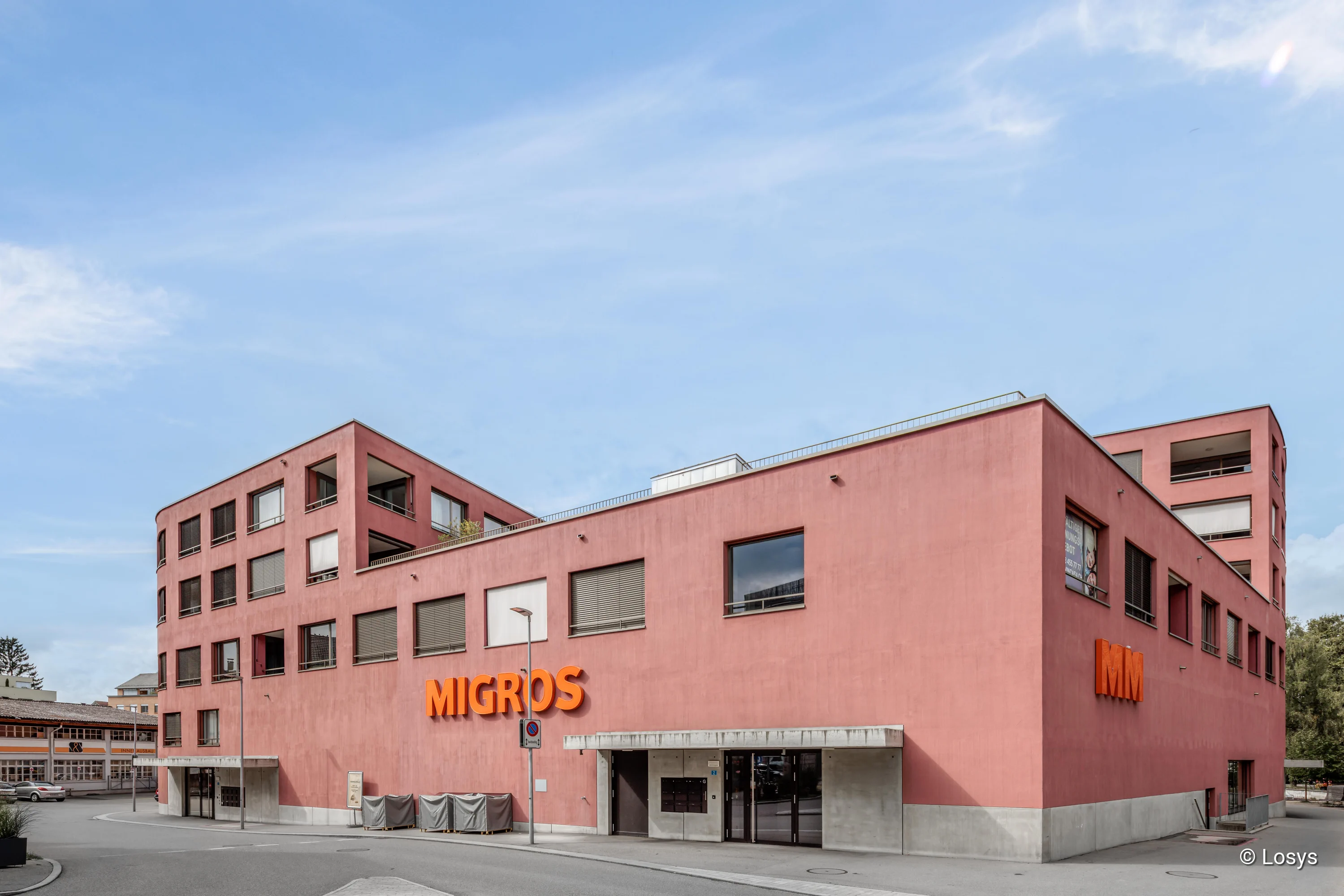 Eckgebäude in rosa-rotem Putz mit orangefarbener MIGROS-Beschriftung, Beton-Sockel und bodentiefem Glas-Eingang (Schiebetür), darüber Wohnungen mit Fensterläden, ruhige Straße im Vordergrund